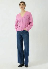 Cardigan rosa tricotado com decote em V, apresentando padrões de corações pretos, combinado com jeans de ganga azul claros com pernas de sino. O design possui mangas longas.