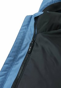 Area della zip della giacca sintetica blu con rivestimento nero, caratterizzata da un'etichetta "ZIP-IN", texture liscia e materiali di tessuto a contrasto.