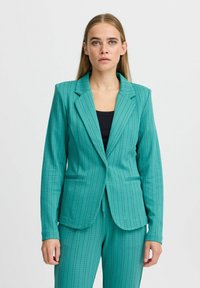 Blazer verde acqua con design a righe, colletto a punta e tasche laterali. Realizzato in tessuto morbido e testurizzato. Silhouette aderente, indossato sopra un top nero.