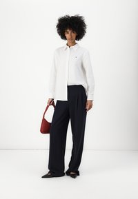 Camisa blanca de botones, pantalones de pierna ancha navy plisados, bolso rojo y zapatos planos negros. Tejido suave; diseño estructurado con un sutil acento de logo.