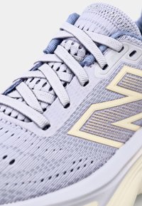 Lys lilla sportsko med et strukturert mesh-overdel, hvite snørebånd, og en gyllen "N"-logo på siden. Merkbare perforeringer for ventilasjon.