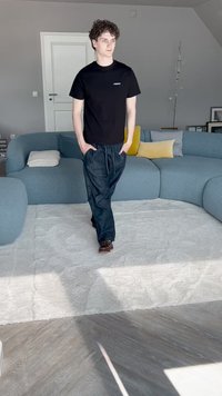 Črn majica s krojem na vratu, ohlapne temno jeans hlače, rjavi vezni čevlji. Sodobna, svetlo siva dnevna soba z modrim kavčem in rumenim akcentom.