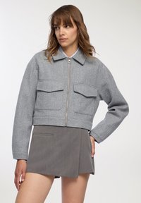 Kobieta w szarej, krótkiej kurtce zapinanej na zamek z dużymi przednimi kieszeniami oraz w szarej mini spódnicy, stojąca z jedną ręką na biodrze.