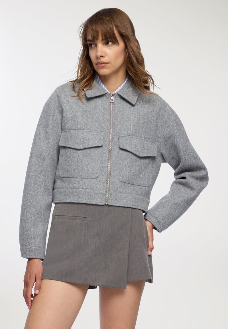 Kobieta w szarej, krótkiej kurtce zapinanej na zamek z dużymi przednimi kieszeniami oraz w szarej mini spódnicy, stojąca z jedną ręką na biodrze.