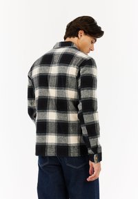 Man in een zwart-wit geruite blouse met lange mouwen en donkerblauwe jeans, zijwaarts gekeerd met het hoofd iets naar beneden, op een witte achtergrond.