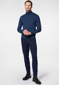 Granatowy sweter z golfem wykonany z miękkiej dzianiny, zestawiony z ciemnymi, dopasowanymi spodniami i czarnymi botkami za kostkę, stojący na jednolitym tle.