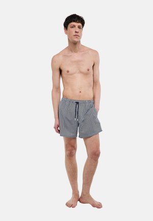 Homme debout pieds nus, une main dans la poche, portant un short de bain à fines rayures noires et blanches avec cordon de serrage, sur un fond uni.