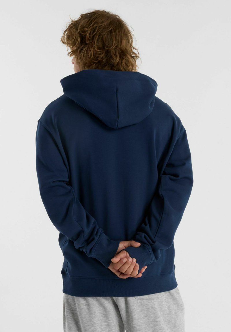 Donkerblauwe hoodie met lange mouwen en een ontspannen pasvorm, voorzien van geribbelde zoom en manchetten, gezien van achteren met in elkaar gevlochten handen.