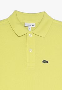 Polo tričko Lacoste v jasně žluté barvě, vyrobené z texturované bavlny s žebrovaným límcem, obsahující dva knoflíky a malé zelené logo na hrudi.