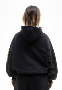 Sudadera corta negra con capucha, que presenta mangas largas y una textura suave. La manga izquierda cuenta con acentos gráficos coloridos.