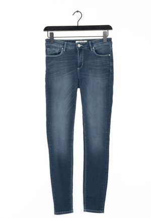 Siniset slim fit -farkut, jotka roikkuvat mustassa henkariassa, etutaskut, nappi ja vyönlenkit näkyvissä valkoista taustaa vasten.