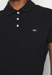 Polo noir à manches courtes avec trois boutons blancs et un petit logo rectangulaire rouge, blanc et bleu sur la poitrine.