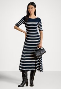 Γυναίκα που φοράει μεσοφόρι φόρεμα με οριζόντιες μπλε σκούρες και λευκές ρίγες, μαύρες μποτάκια μέχρι το γόνατο, κρατώντας μαύρη τσάντα με λεπτομέρειες αγκράφας.