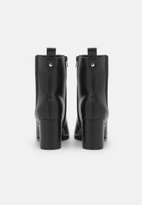 Par de botas de cuero negro hasta el tobillo con tacones cuadrados vistas desde atrás sobre un fondo blanco.