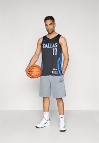 Schwarzes Basketballtrikot mit "DALLAS" und der Nummer "11" in Blau, graue Shorts, hält einen orangenen Basketball, trägt weiße Nike-Schuhe.