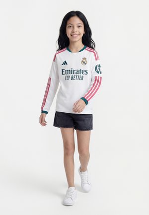 Dívka kráčí vpřed, má na sobě bílý dlouhý rukáv dres Realu Madrid, černé kraťasy a bílé tenisky, usmívá se na jednobarevném pozadí.