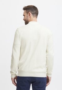 Lyscreme strikket sweater med et struktureret mønster, ribbede manchetter og søm, samt en rund krave. Bagsiden viser detaljer ved nakken og ærmerne.