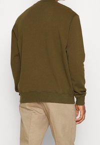 Sudadera verde oliva con cuello redondo, mangas largas, puños y dobladillo acanalados, fabricada en un tejido suave, llevada sobre pantalones beige claros.