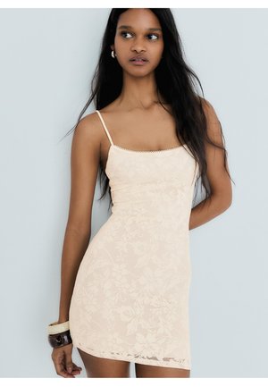 Vrouw met lang donker haar die een aansluitende crèmekleurige kanten jurk met dunne bandjes draagt, staand tegen een lichtgrijze achtergrond.