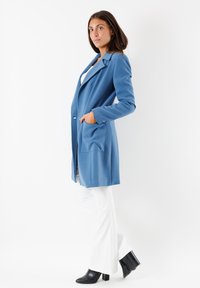 Cappotto blu su misura con rever a notch, due tasche frontali e una texture liscia, abbinato a pantaloni bianchi svasati e stivaletti neri alla caviglia.