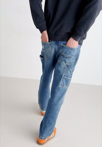 Light blue denim cargo pants with multiple pockets and a relaxed fit, paired with orange and cream-striped sneakers. Casual style.