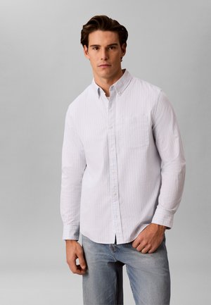 Mladý muž nosiaci bielu košeľu s tenkými vertikálnymi pruhmi a svetlomodré rifle, stojaci pred jednoduchým sivým pozadím.