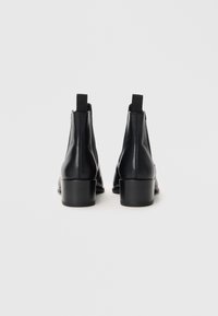 Botas de tornozelo em couro preto com um salto quadrado baixo e painéis laterais elásticos. Textura suave com detalhes de costura mínimos, apresentando alças de puxar.