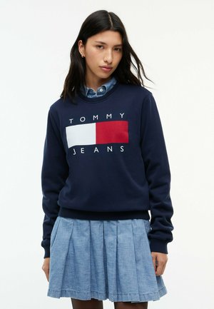 Kobieta nosi granatową bluzę Tommy Jeans na niebieskiej koszuli z guzikami oraz jasnoniebieską plisowaną spódnicę.