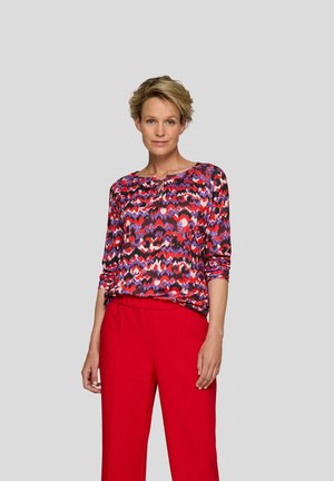 Multicolored patterned top in shades of purple, red, and white, with a round neckline and three-quarter sleeves, paired with red pants.