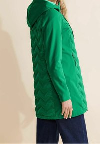 Groene capuchonjas met een doorgestikt chevronpatroon aan de zijkant. Voorzien van een gladde stof, ritssluiting aan de voorkant en lange mouwen.