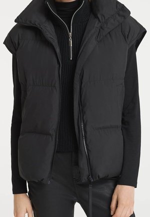 Schwarze ärmellose Steppweste mit Reißverschluss, getragen über einem schwarzen, gerippten Pullover mit halbem Reißverschluss und schwarzen Hosen, gezeigt vom Hals bis zur Hüfte.