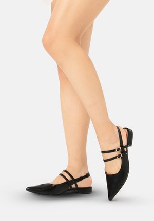 Benen met zwarte platte slingbackpumps met puntige neus, dubbele dunne bandjes en kleine gouden gespen tegen een effen lichte achtergrond.