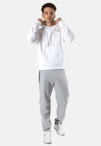 Sudadera blanca con capucha y logo dorado, de manga larga y con bolsillo delantero, combinada con pantalones de chándal grises que tienen bolsillos con cremallera y puños elásticos.