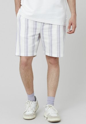 Shorts rayés en blanc avec des lignes lavande, fabriqués à partir d'un tissu léger. Coupe décontractée, poches latérales, portés avec des baskets blanches et des chaussettes lavande.