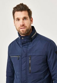 Navy blue insulated jacket featuring a high collar, zippered front and chest pockets, and contrasting white stitching details.