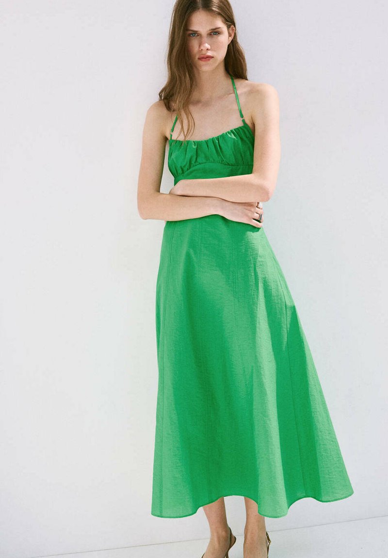 Jeune femme aux longs cheveux portant une robe maxi verte sans manches, debout les bras croisés devant un fond blanc uni.