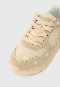 Baskets beige avec des lacets épais, des accents en daim, et de petits motifs floraux brodés en blanc, jaune et bleu sur l'avant et les côtés.