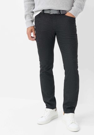 Mand iført mørkegrå bukser, en grå sweater, hvide sneakers og et mørkt bælte, stående med den ene hånd i lommen mod en ensfarvet baggrund.
