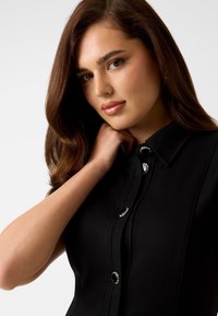 Černé šaty s límečkem, zdobené stříbrnými knoflíky a výrazným prošíváním. Hladká textura a přizpůsobený střih.