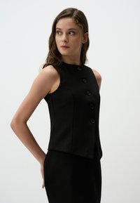 Gilet nero senza maniche con sei bottoni frontali, vestibilità sagomata, texture liscia e scollatura alta. Indossato sopra un vestito nero coordinato.