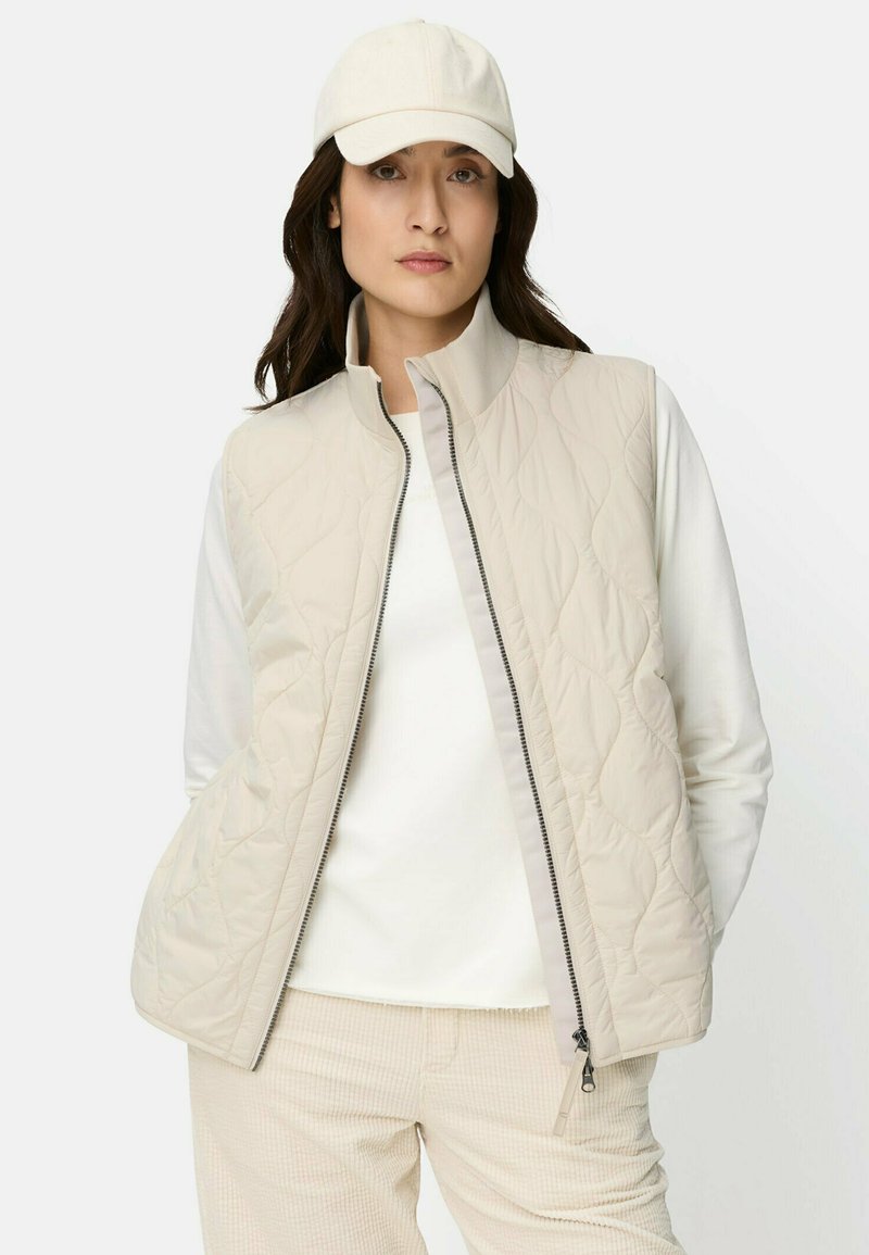 Hellbeige gesteppte Weste mit hohem Kragen und Reißverschluss, getragen über einem weißen Langarmshirt, kombiniert mit cremefarbenen Hosen.