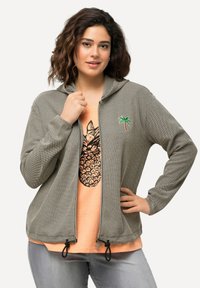 Lys grå zip-up hoodie med et struktureret mønster, prydet med en broderet palmetræ. Båret over en koralfarvet t-shirt med et ananasgrafik.