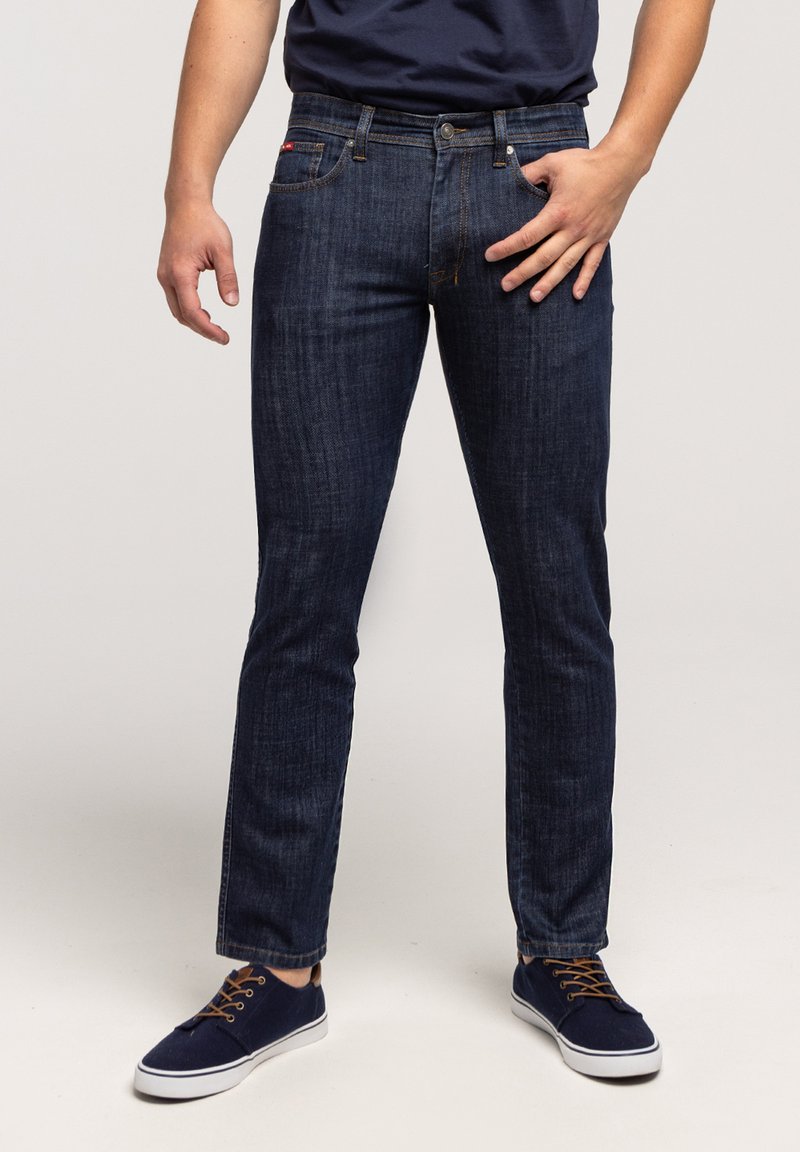 Mężczyzna ubrany w ciemnoniebieskie, obcisłe dżinsy i granatowe trampki z brązowymi sznurowadłami, stojący na tle jednolitego jasnego tła.