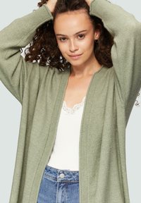 Hellgrüner Strickcardigan mit offenem Front und gerippten Bündchen, getragen über einem weißen, mit Spitze verzierten Tanktop und blauen High-Waist-Jeans.