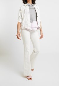 Giacca di jeans bianca sopra un top grigio, abbinata a jeans bianchi svasati e sandali rosa; texture liscia, design casual a strati e colori chiari.