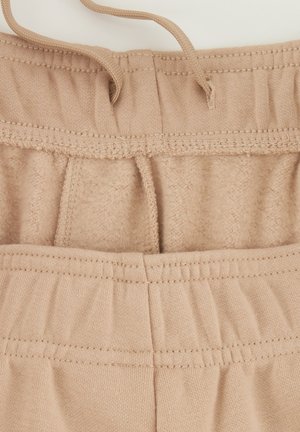 Un pantalon de survêtement beige avec un intérieur doux, une taille élastique et un cordon de serrage. Le tissu a une texture lisse avec des détails de couture visibles.