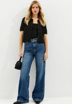 Mujer joven con cárdigan negro de manga corta abotonado, top negro, vaqueros azules de pierna ancha, zapatos negros, sosteniendo un bolso pequeño negro.