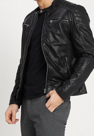 Giubbotto motociclista in pelle nera con tasche con zip, spalle trapuntate e design aderente. Abbinato a una maglietta nera e pantaloni grigi con texture.