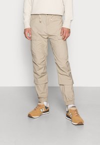 Muž v béžových přestavitelných cargo kalhotách s elastickými manžetami a hnědých teniskách New Balance, stojící proti bílému pozadí.
