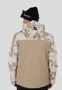 Beige-weiße camouflage Jacke mit Kapuze, mit einem Logo auf dem Rücken und einer glatten Textur. Getragen mit einer schwarzen Mütze und Skibrille.